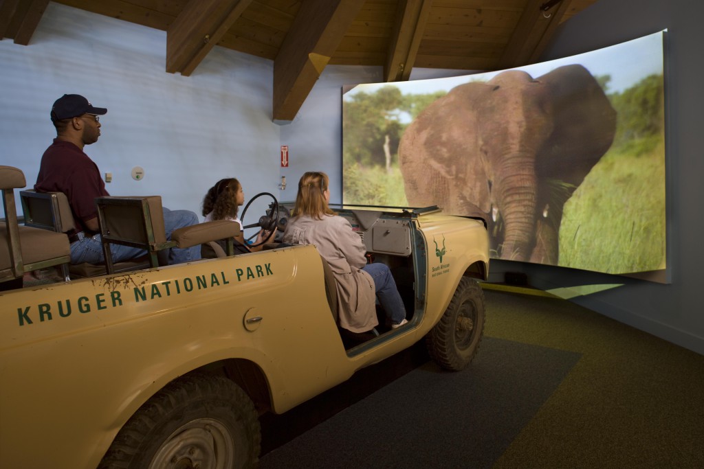 Three people on a jeep with an elephant projected onto a screen in front of them